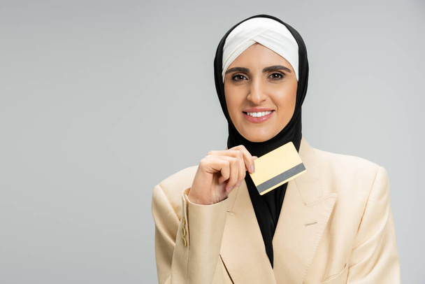 mujer de negocios musulmana con estilo en blazer y hijab celebración de la tarjeta de crédito y sonriendo en gris, headshot - Foto, imagen