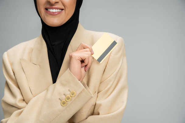 灰色のクレジットカードを持っているブレザーとヒジャブで笑顔のイスラム教徒のビジネスマンのビュー - 写真・画像