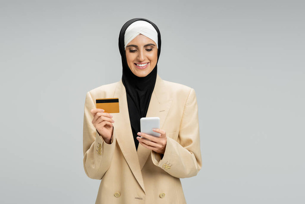 Muslimische Geschäftsfrau im Hijab und schickem Blazer hält Smartphone und Kreditkarte auf grau - Foto, Bild