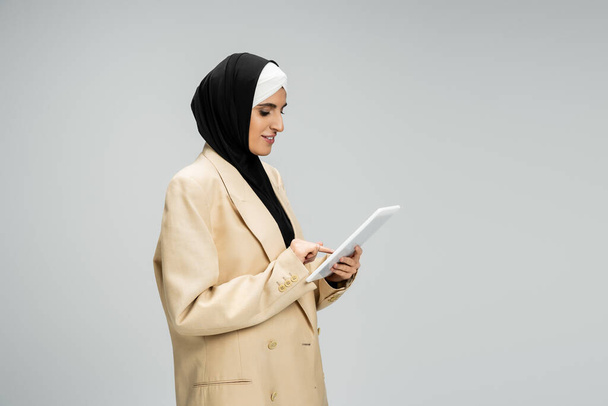 mujer de negocios musulmana positiva en hijab y beige blazer networking en tableta digital en gris - Foto, imagen