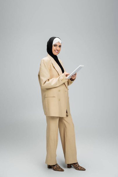 šťastná muslimská obchodnice v béžovém obleku a hidžábu stojící s digitálním tabletem na šedé, plné délce - Fotografie, Obrázek