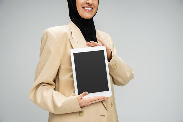 oříznutý pohled usměvavé muslimské podnikatelky držící digitální tablet s prázdnou obrazovkou na šedé - Fotografie, Obrázek