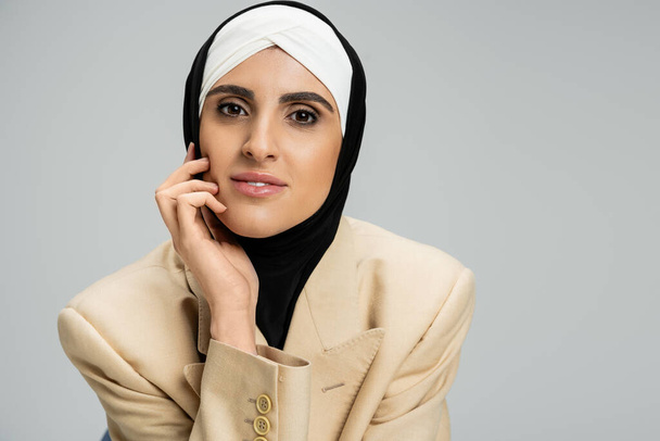portrait of modern and trendy muslim businesswoman in hijab and blazer looking at camera on grey - Zdjęcie, obraz