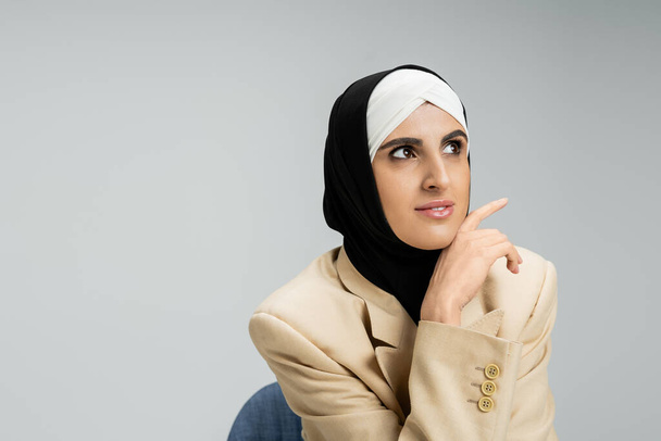 mujer de negocios musulmana de moda en hijab y blazer beige sentado y mirando hacia otro lado en gris - Foto, Imagen