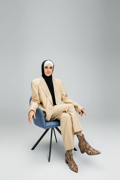ambiciosa mujer de negocios musulmana en traje de moda y hijab sentado en sillón en gris, longitud completa - Foto, Imagen