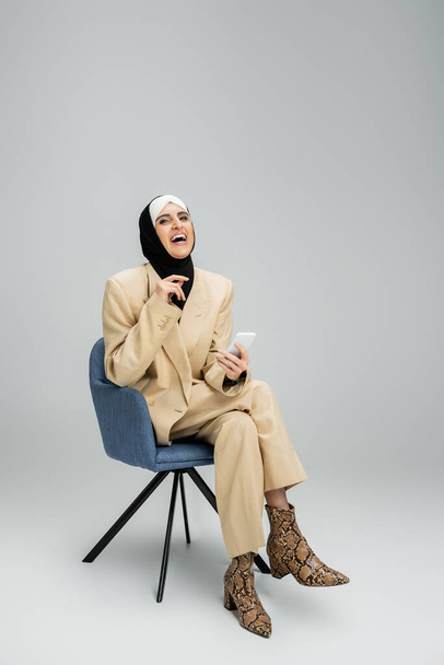 mujer de negocios musulmana emocionada y con estilo con teléfono inteligente sentado en sillón y riendo en gris - Foto, Imagen