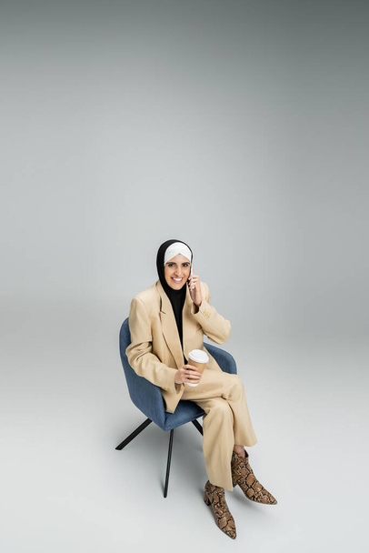 alegre, mulher de negócios muçulmana na moda com café para ir falar no smartphone em poltrona em cinza - Foto, Imagem