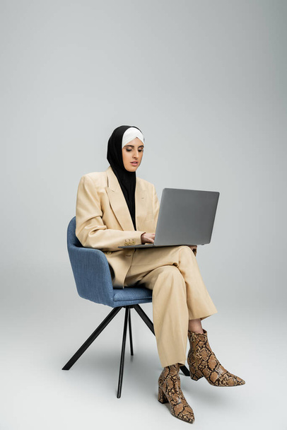 Kafiyeli ve takım elbiseli modern Müslüman iş kadını gri koltukta, tam boy dizüstü bilgisayarda çalışıyor. - Fotoğraf, Görsel