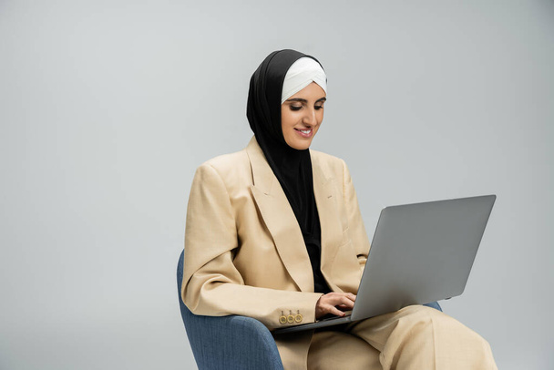 ευχαριστημένη, μοντέρνα μουσουλμάνα επιχειρηματίας με μαντίλα και μπεζ κοστούμι που εργάζεται σε φορητό υπολογιστή σε πολυθρόνα σε γκρι - Φωτογραφία, εικόνα