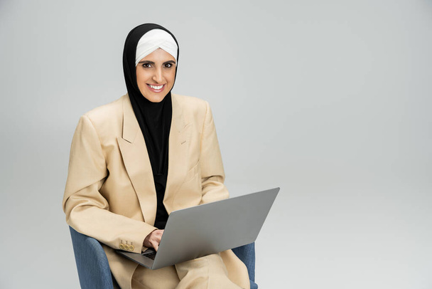 odnosząca sukcesy i modna muzułmańska bizneswoman z laptopem uśmiechnięta do kamery w fotelu na szarości - Zdjęcie, obraz