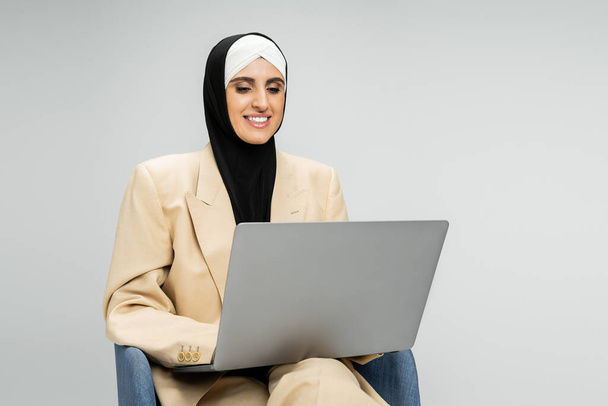 tevreden zakenvrouw in pak en hijab netwerken op laptop in fauteuil op grijs - Foto, afbeelding