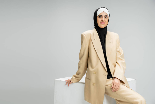exitosa mujer de negocios musulmana en traje elegante y hijab sonriendo a la cámara cerca del cubo blanco en gris - Foto, imagen