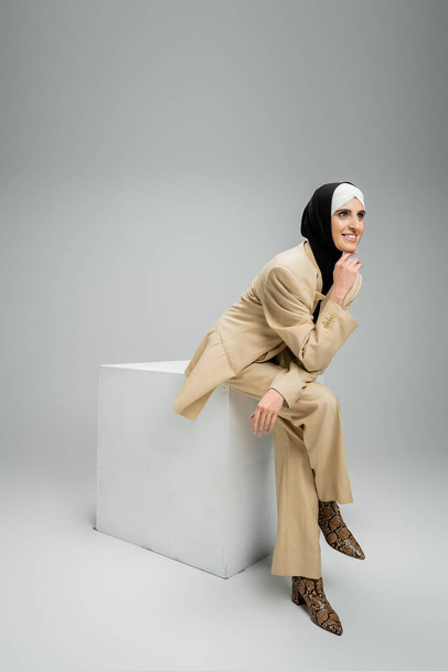 mujer de negocios musulmana sonriente en hijab y elegante traje mirando hacia otro lado en cubo en gris, longitud completa - Foto, Imagen