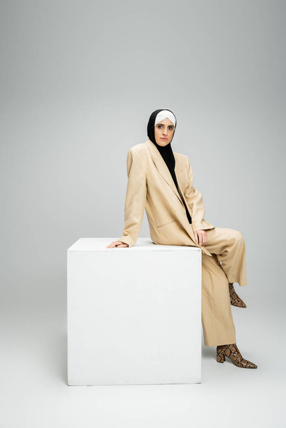 уверенная мусульманка-бизнесмен в бежевом стильном костюме и хиджабе, сидящая на белом кубе на сером - Фото, изображение