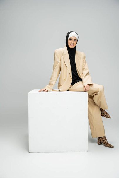 cheerful muslim businesswoman in fashionable attire and hijab sitting on white cube on grey - Photo, Image