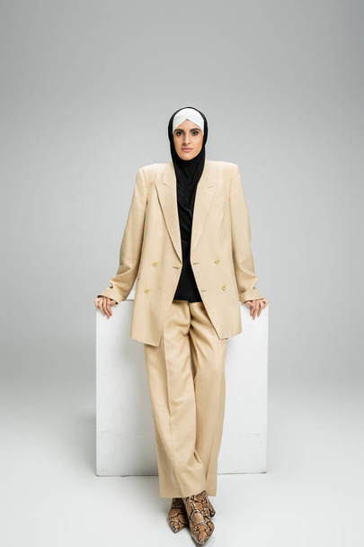 onafhankelijke moslim zakenvrouw in stijlvolle pak en hijab staan in de buurt kubus op grijs, volledige lengte - Foto, afbeelding