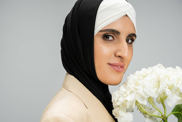 Porträt einer eleganten muslimischen Geschäftsfrau mit weißen Hortensienblüten, die in die Kamera auf grau blickt - Foto, Bild