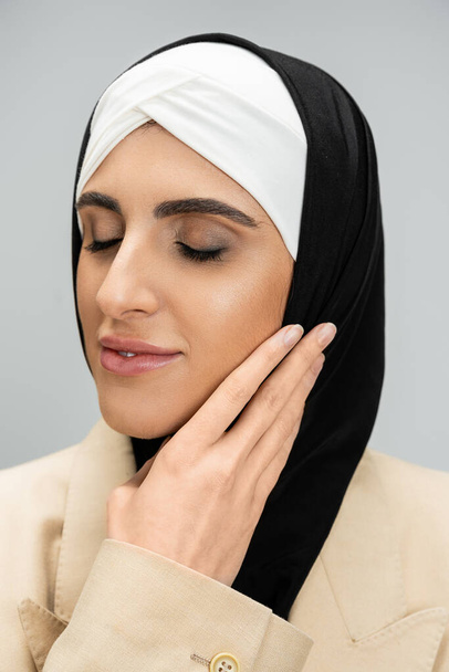 retrato de mujer de negocios musulmana encantada y elegante con los ojos cerrados tocando la cara en gris - Foto, imagen
