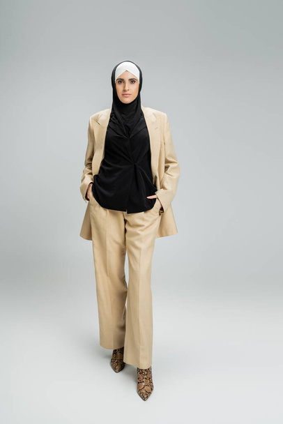 empresaria musulmana contemporánea en traje beige y hijab posando con las manos en bolsillos en gris - Foto, imagen