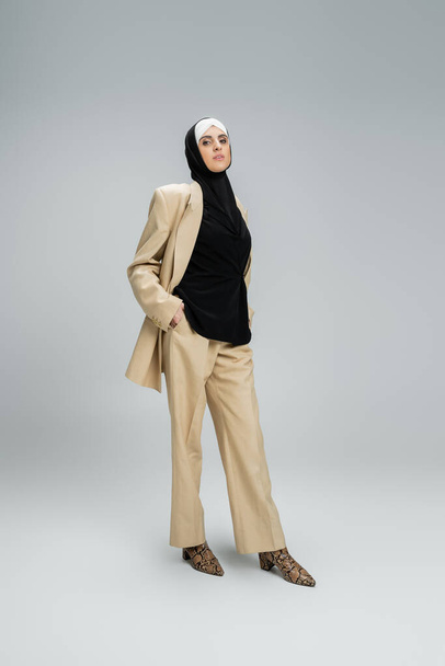 zelfverzekerde muslin vrouw in hijab en stijlvolle zakelijke kleding op zoek naar camera op grijs, volledige lengte - Foto, afbeelding