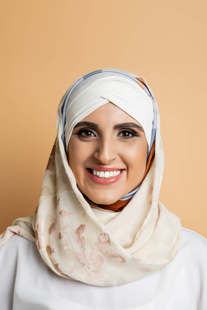 portret czarującej muzułmanki z makijażem, w jedwabnym szaliku na głowie uśmiechnięty do aparatu na beżu - Zdjęcie, obraz