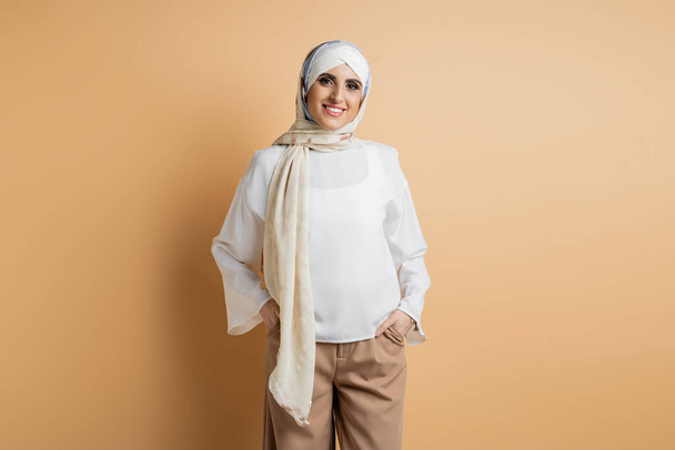 fröhliche Muslimin in weißer Bluse und seidenem Kopftuch mit Händen in Taschen auf beige - Foto, Bild