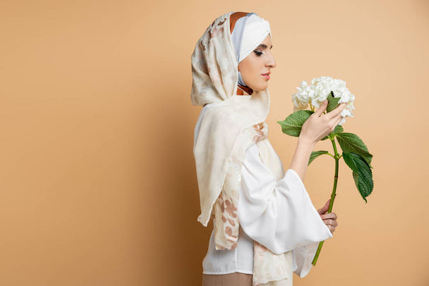 anmutige muslimische Frau in seidenem Kopftuch und Bluse mit weißer Blume auf beige, Seitenansicht - Foto, Bild