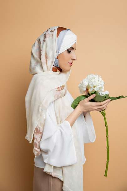 vista lateral de la mujer musulmana de moda en pañuelo para la cabeza y blusa blanca con flor de hortensia en beige - Foto, imagen