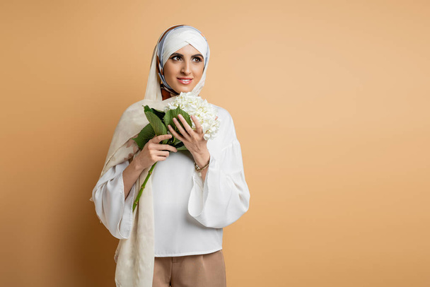 schöne muslimische Frau in seidenem Kopftuch und Bluse stehend mit weißen Hortensienblüten auf beige - Foto, Bild