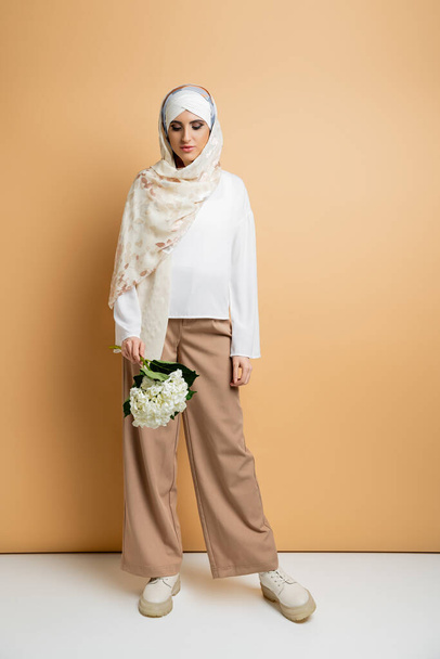 donna musulmana moderna in fazzoletto di seta ed elegante abbigliamento casual in posa con fiore bianco su beige - Foto, immagini