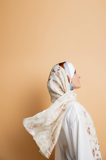 πλαϊνή όψη του χαμογελαστού μουσουλμάνου γυναίκα σε κομψό μεταξωτή μαντίλα και λευκή μπλούζα σε μπεζ - Φωτογραφία, εικόνα