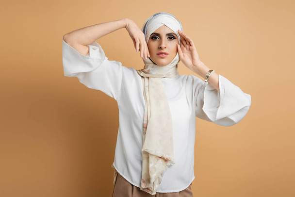 elegante donna musulmana in camicetta bianca e velo di seta posa con le mani vicino al viso sul beige - Foto, immagini