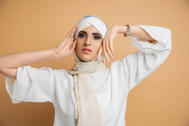 graceful muslim woman in elegant attire holding hands near face and looking at camera on beige - Photo, Image
