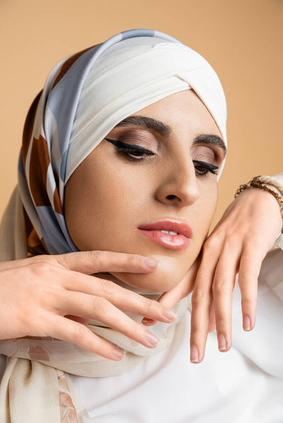 πορτρέτο της σύγχρονης γυναίκας μουσουλμάνος με μακιγιάζ και τα χέρια κοντά στο πρόσωπο ποζάρουν σε μεταξωτή μαντίλα σε μπεζ - Φωτογραφία, εικόνα
