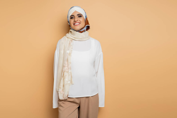 allegra donna musulmana in camicetta bianca e sciarpa di seta in piedi e sorridente alla macchina fotografica sul beige - Foto, immagini