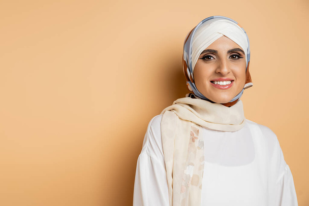 gelukkig moslim vrouw met make-up dragen zijden sjaal en witte blouse, glimlachen op camera op beige - Foto, afbeelding