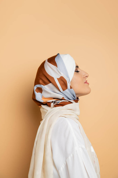 Profil der attraktiven Muslimin in elegantem Seidenkopftuch, die mit geschlossenen Augen auf beige posiert - Foto, Bild