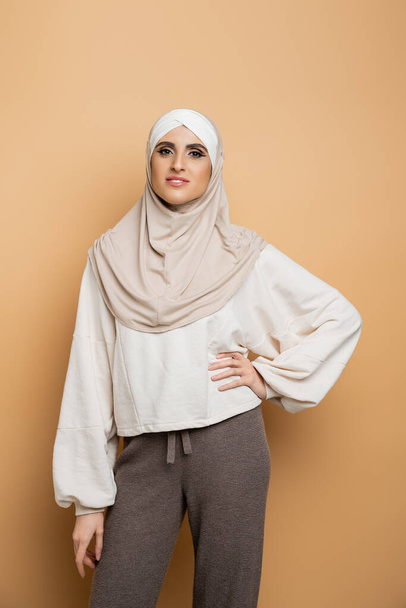 gelukkig moslim vrouw in hijab en stijlvolle casual kleding poseren met de hand op heup op beige - Foto, afbeelding
