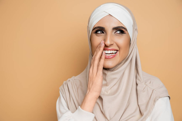excited, stylish muslim woman in hijab covering mouth with hand, looking away and laughing on beige - Fotó, kép