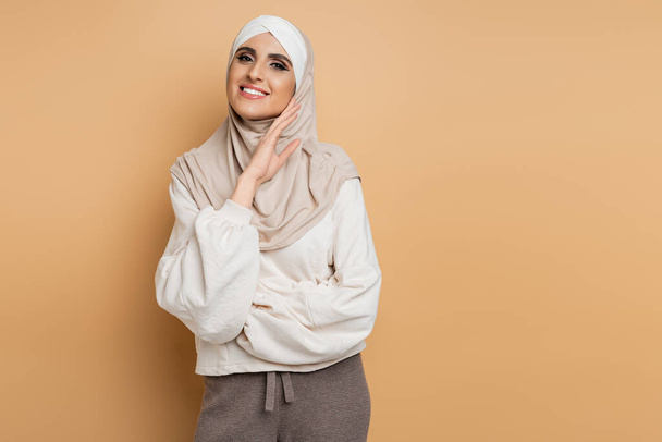 happy muslim woman in hijab and trendy casual attire looking at camera with hand near face on beige - Photo, Image