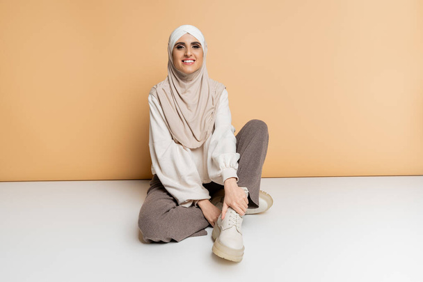 mujer musulmana feliz en hijab y atuendo casual moderno sentado en beige, longitud completa, estilo personal - Foto, Imagen