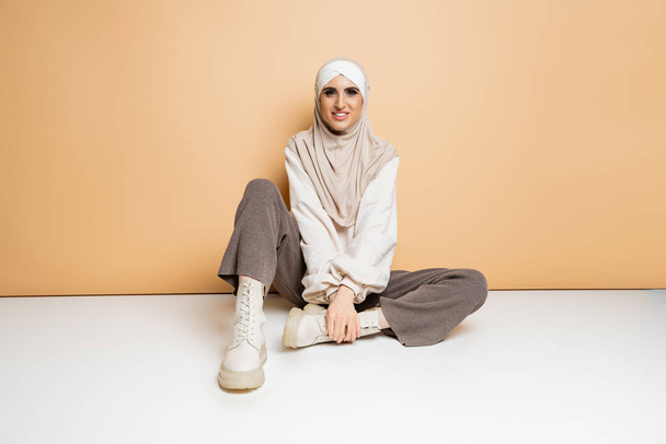 χαρούμενη μουσουλμάνα γυναίκα με μαντίλα, παντελόνι και δερμάτινες μπότες κάθεται και κοιτάζει την κάμερα σε μπεζ - Φωτογραφία, εικόνα