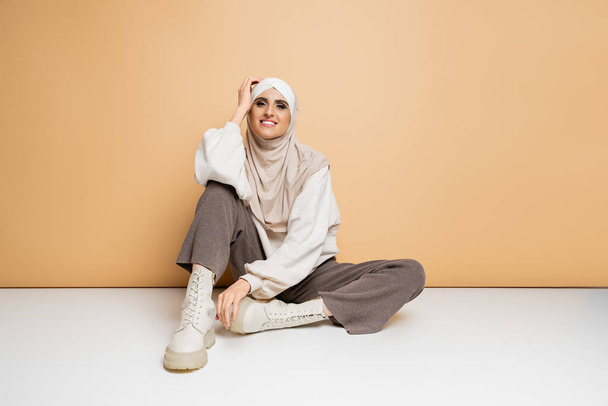 mujer musulmana de moda en hijab, sudadera, pantalones y botas sentados y sonriendo a la cámara en beige - Foto, Imagen