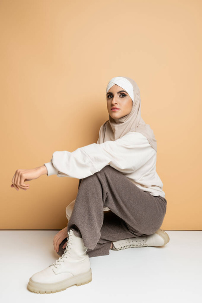 mujer musulmana confiada en hijab, pantalones grises y botas de cuero sentado y mirando a la cámara en beige - Foto, imagen