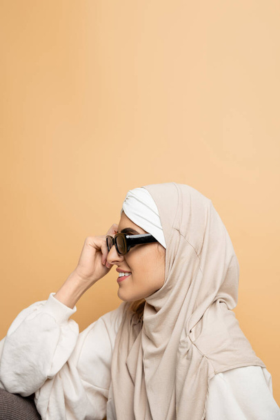 side view of muslim woman in stylish sunglasses and hijab smiling on beige, modern individuality - Fotografie, Obrázek