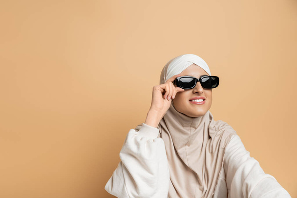 uśmiechnięta muzułmanka w hidżabie i modne okulary przeciwsłoneczne odwracające wzrok na beżowej, nowoczesnej autoekspresji - Zdjęcie, obraz