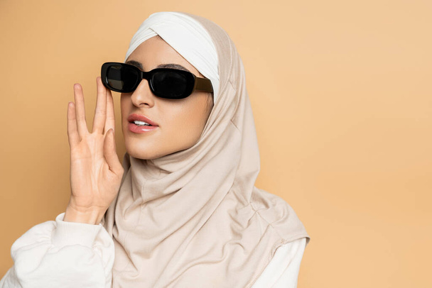 portrait of modern muslim woman in hijab adjusting trendy sunglasses and looking away on beige - Photo, Image