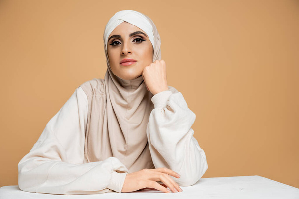 fashionable muslim woman in hijab and sweatshirt sitting at table, looking at camera woman on beige - Foto, Imagem