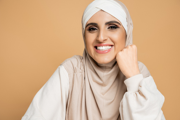 χαρούμενη μουσουλμάνα γυναίκα με χιτζάμπ και λευκό φούτερ χαμογελώντας στην κάμερα σε μπεζ, μοντέρνο τρόπο ζωής - Φωτογραφία, εικόνα