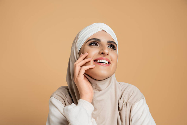 excited muslim woman in white sweatshirt and hijab holding hand near face and looking away on beige - Photo, Image